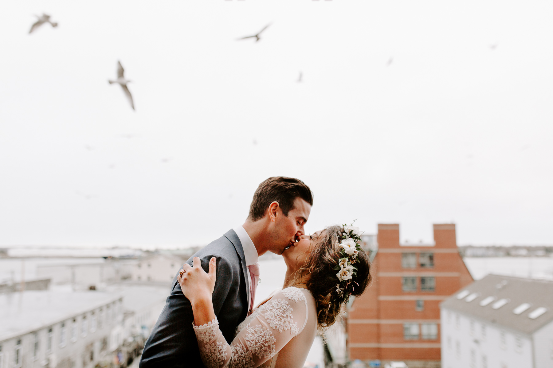 A bride and groom kiss overlooking Portland Harbor in Portland, Maine and sea birds fly above