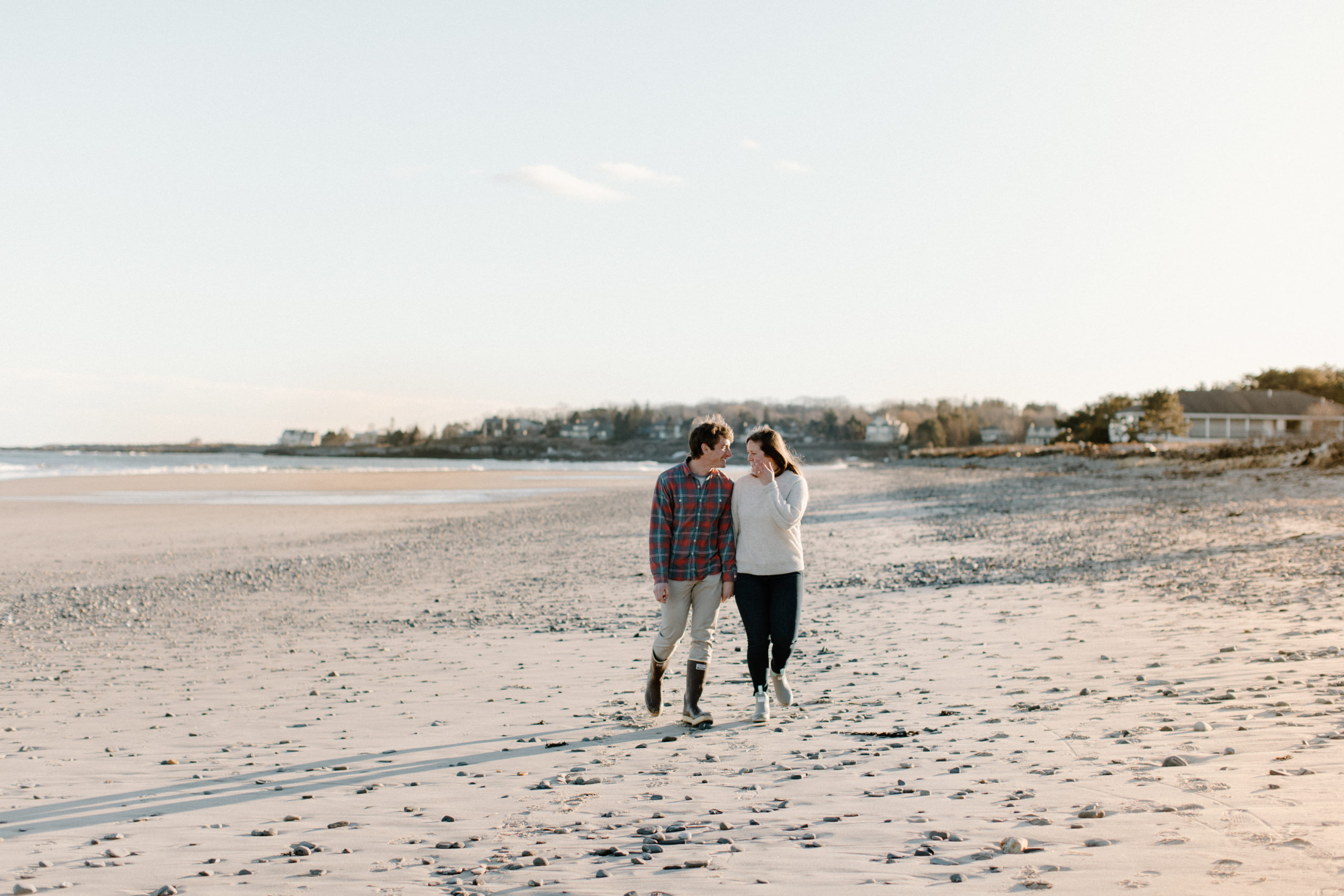 An engaged couple looks at one another while walking down the beach in Maine